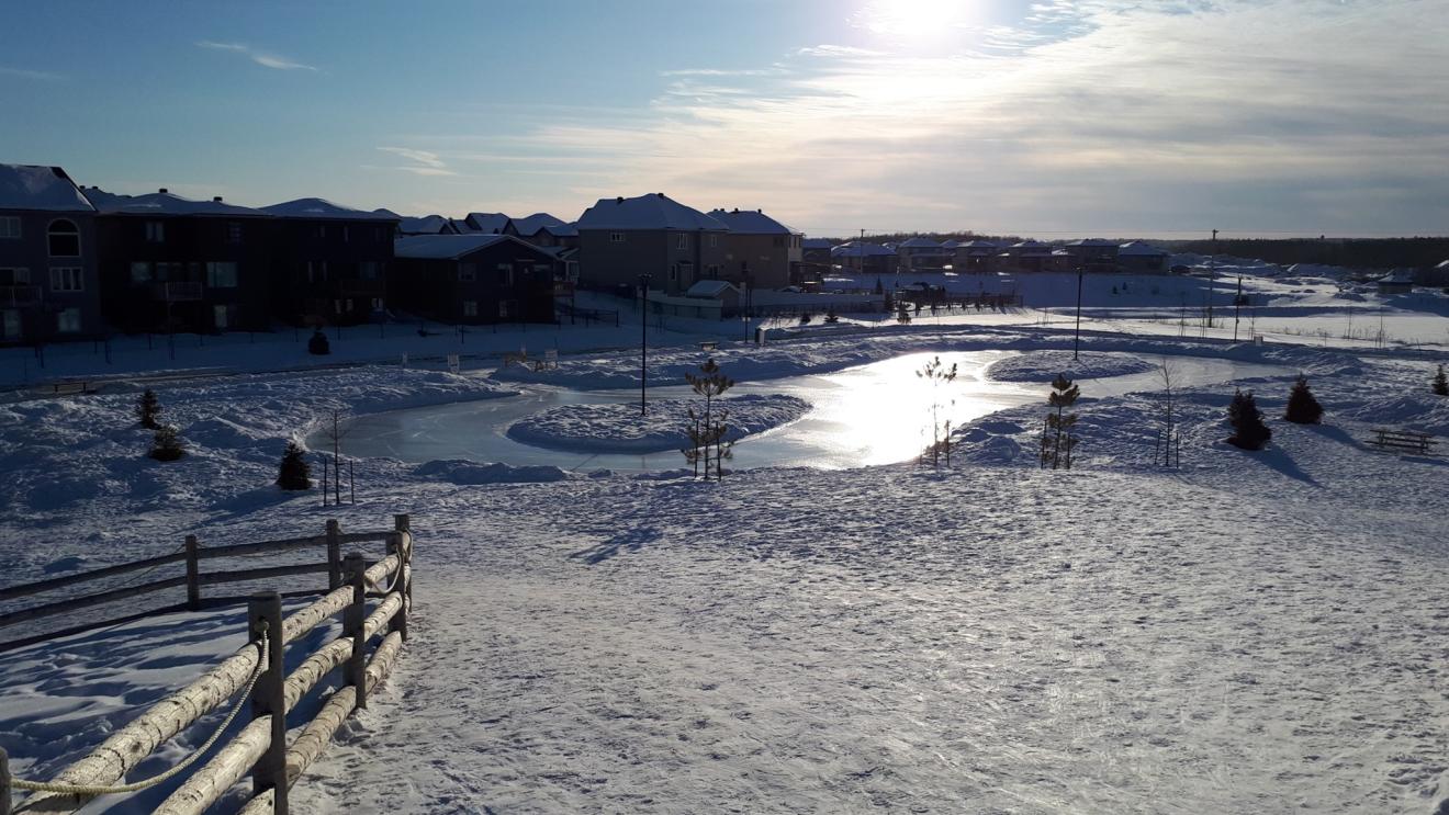 Snowy landscape with a frozen pond and houses in the distance under a bright sun.