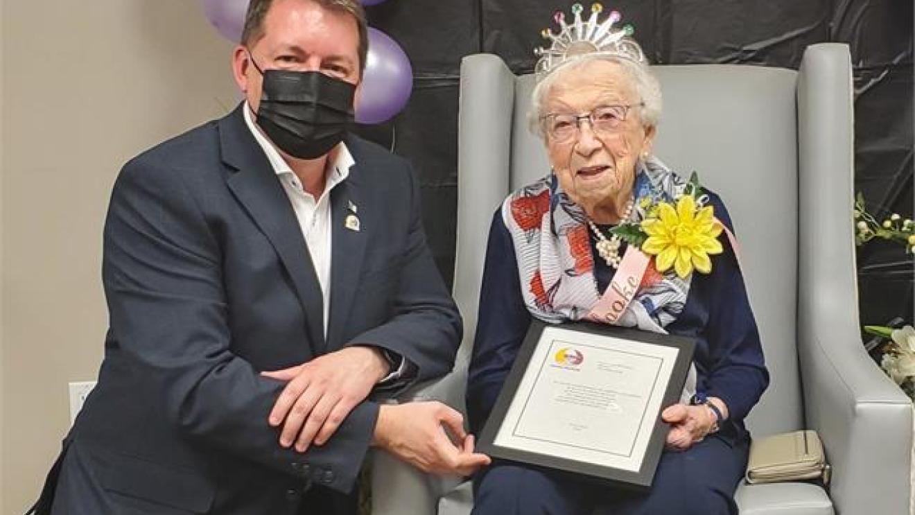 Elderly woman with crown holds certificate, seated next to a masked man, balloons in background.