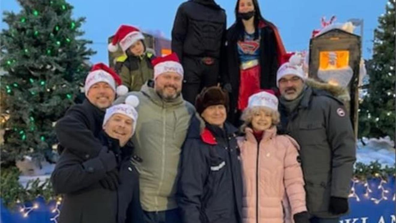 Group in winter clothing and Santa hats posing outdoors near Christmas trees.