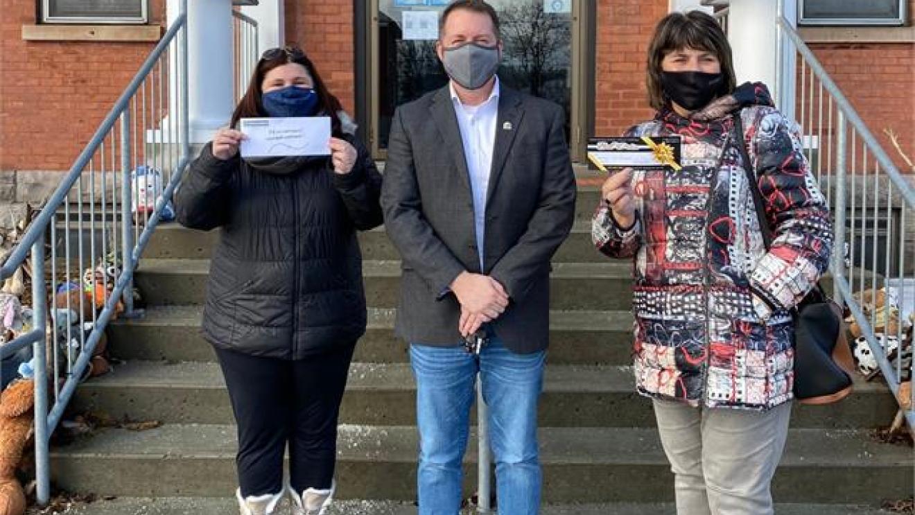 Three people in masks stand on steps holding papers, smiling.