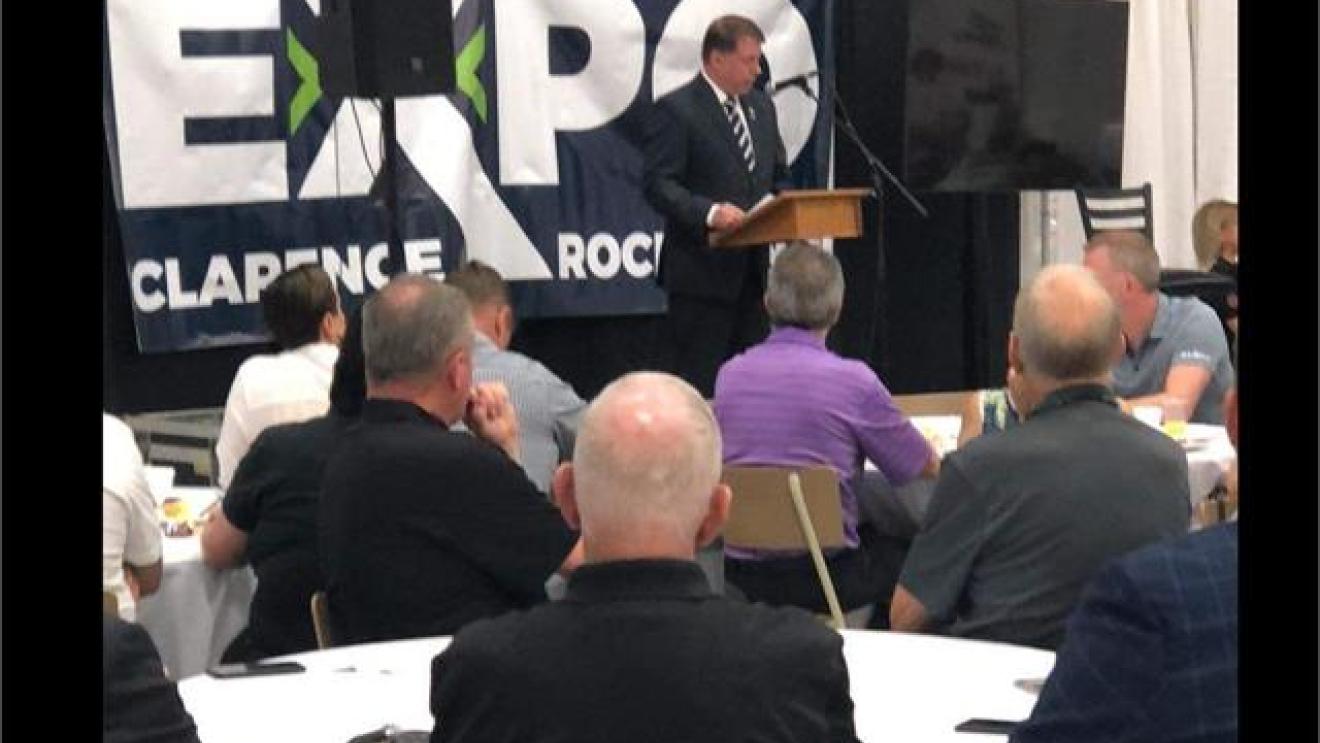 Man speaking at a podium, audience seated at round tables. Conference setting.