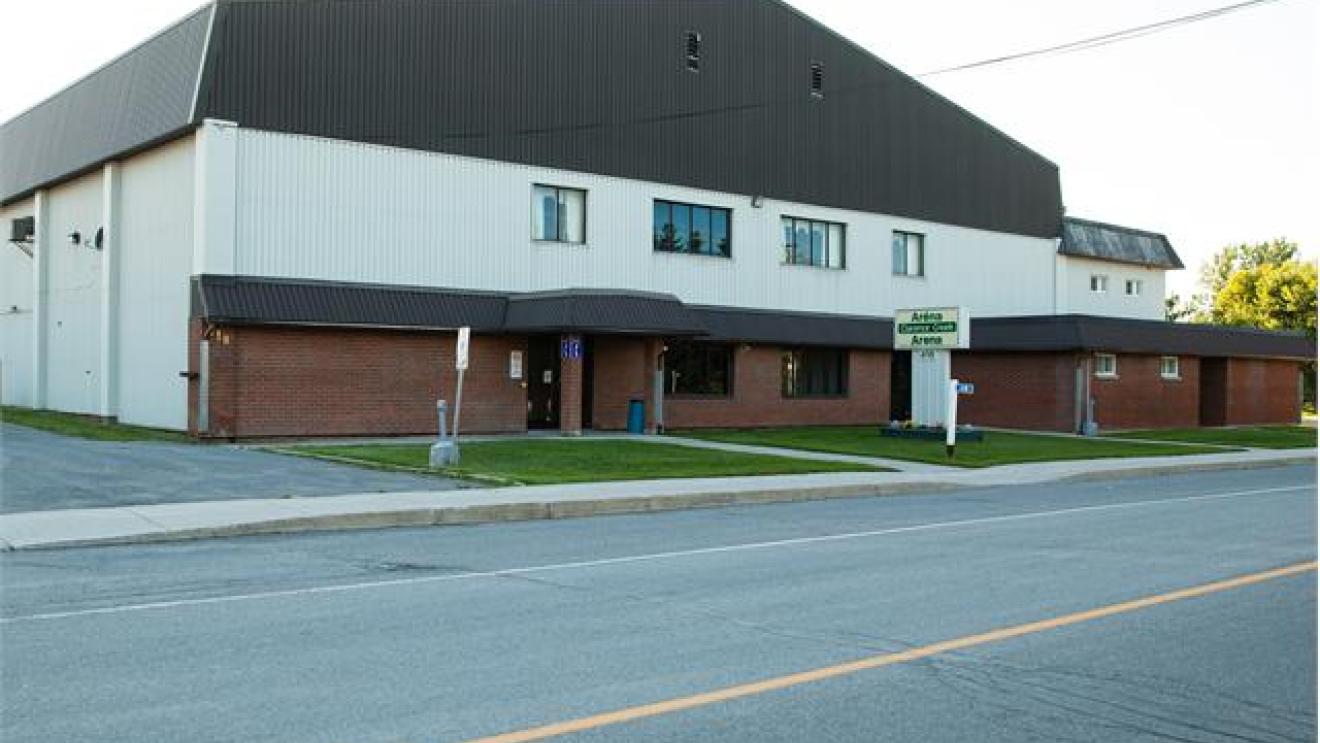 Large tan building with dark roof and a parking area in front.