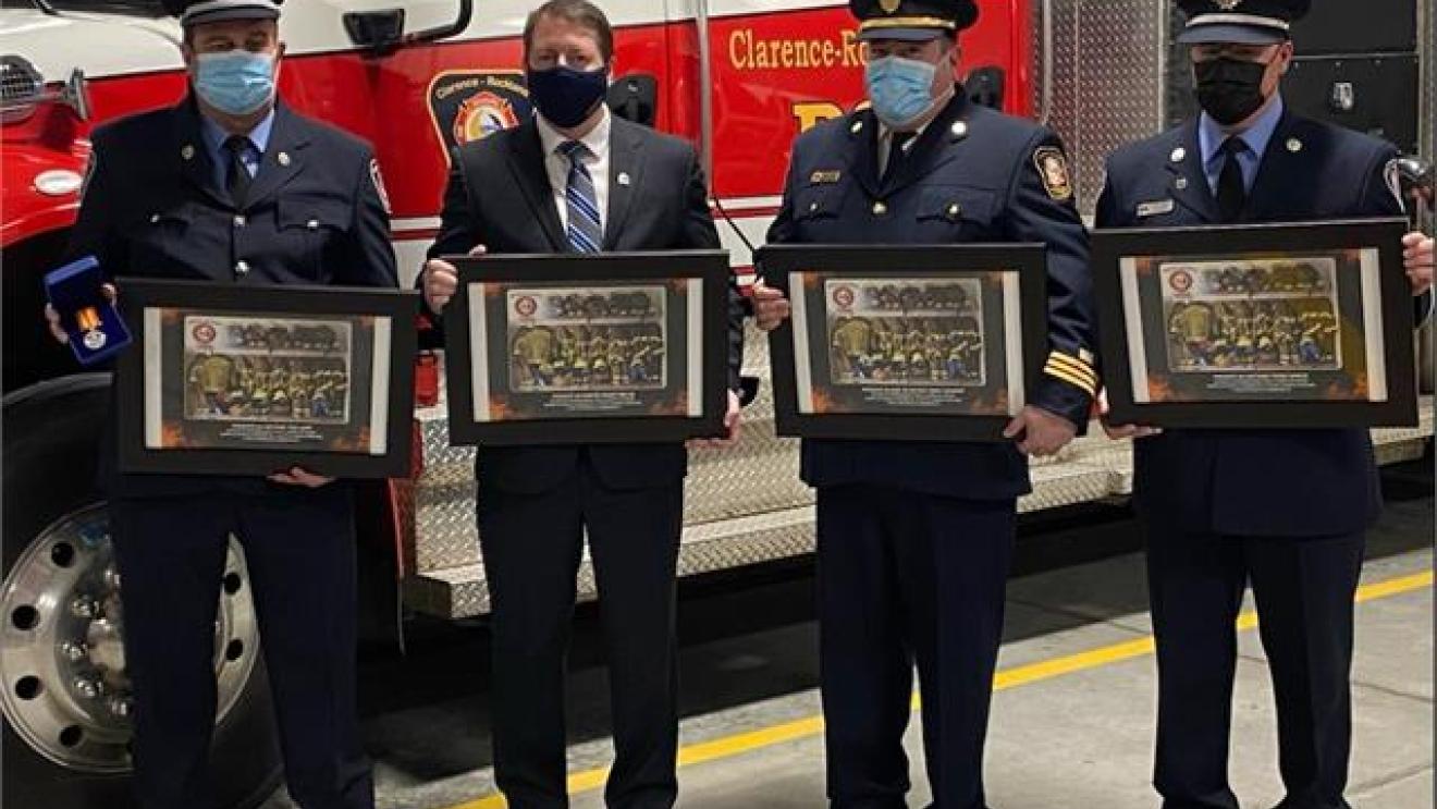 Four uniformed firefighters holding framed awards stand in front of a fire truck.