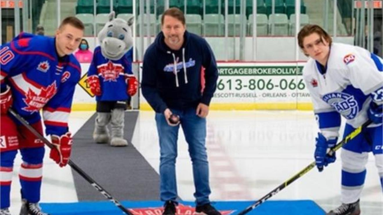 Hockey players face off at center ice with a man dropping the puck, mascot in background.