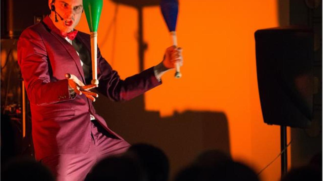 Performer juggling clubs on stage, wearing a purple suit, with orange backdrop.