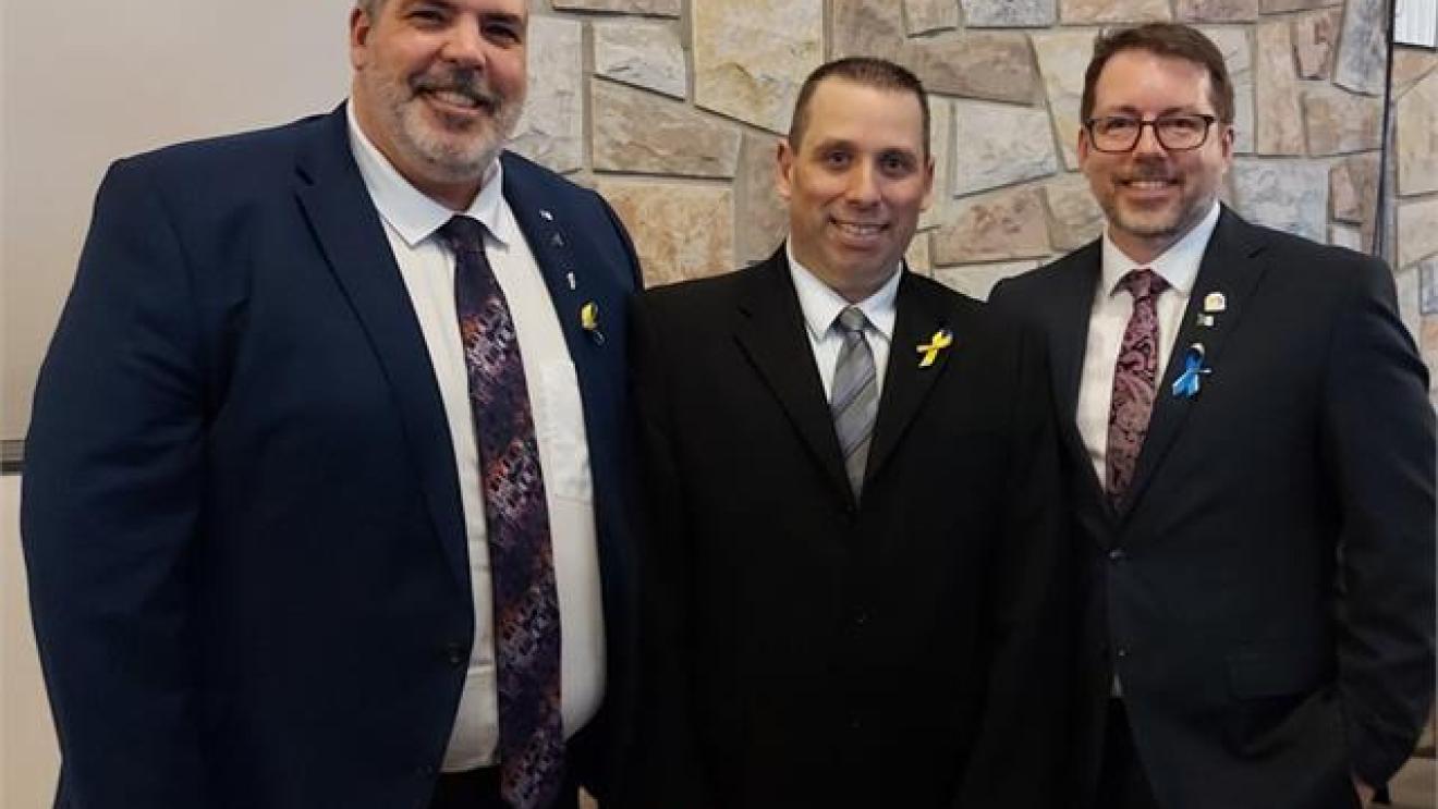 Three men in suits smiling indoors against a stone wall.