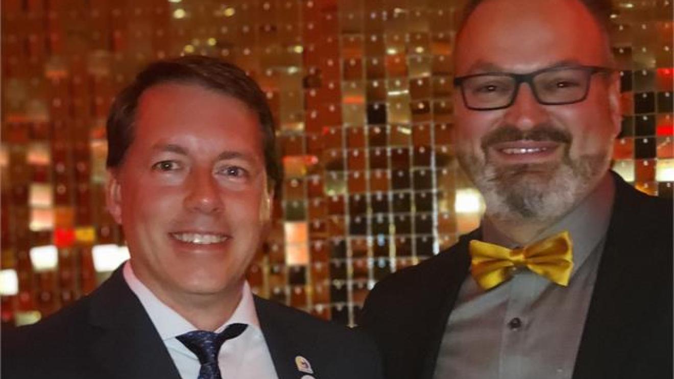 Two men in formal wear smiling, one with glasses and a yellow bow tie, in a warm-lit setting.