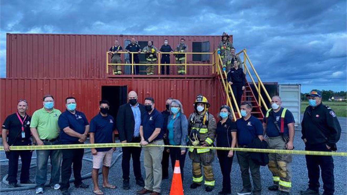 Group of people, some in uniforms, stand by shipping containers under a cloudy sky.