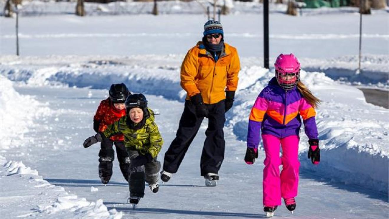 Children and an adult ice skating on a snowy path, wearing colorful winter clothes.