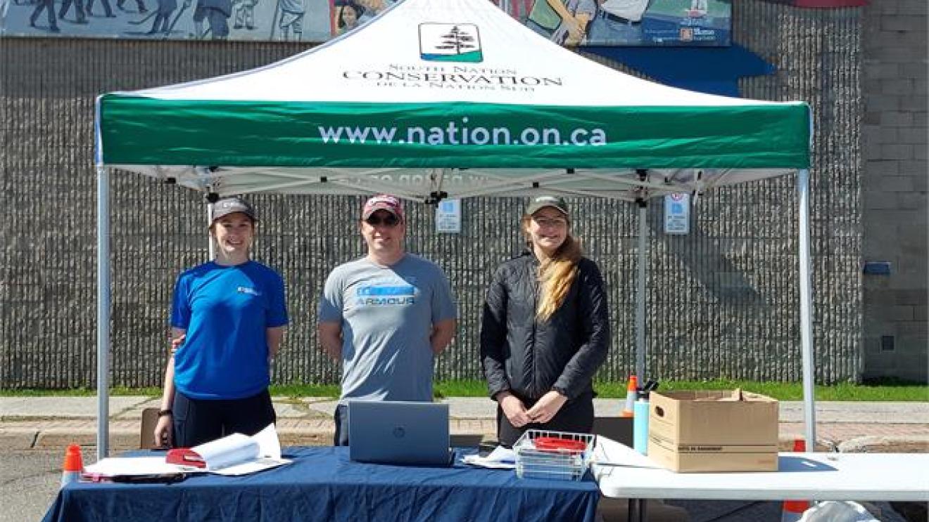 Three people stand under a tent at an outdoor event, smiling.