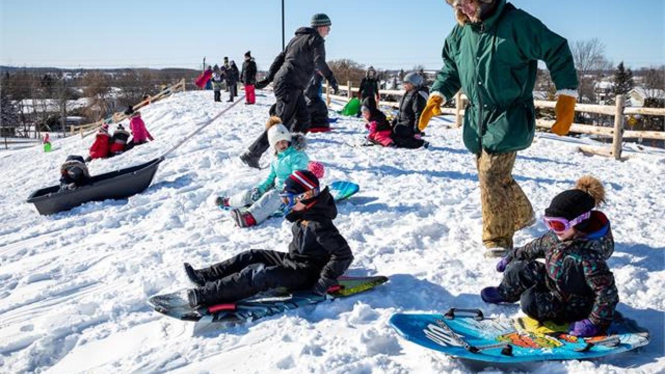 Children sledding down a snowy hill with an adult nearby.