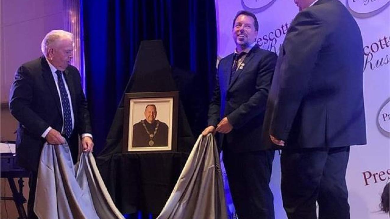 Three men unveil a portrait on a stage with purple lighting.