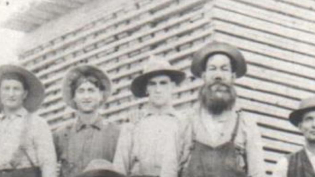 Group of people in hats standing in front of a wooden building, sepia-toned photograph.