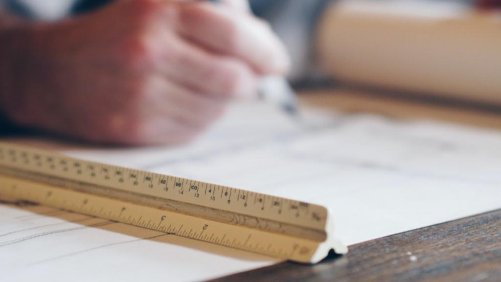 Person writing on architectural plans with a ruler on a table.