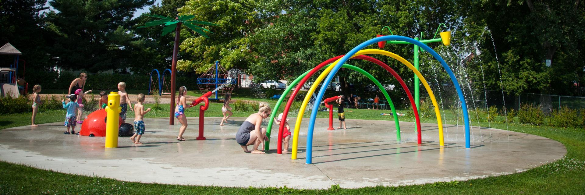 Children play in a colourful splash pad with arched sprinklers in a park.