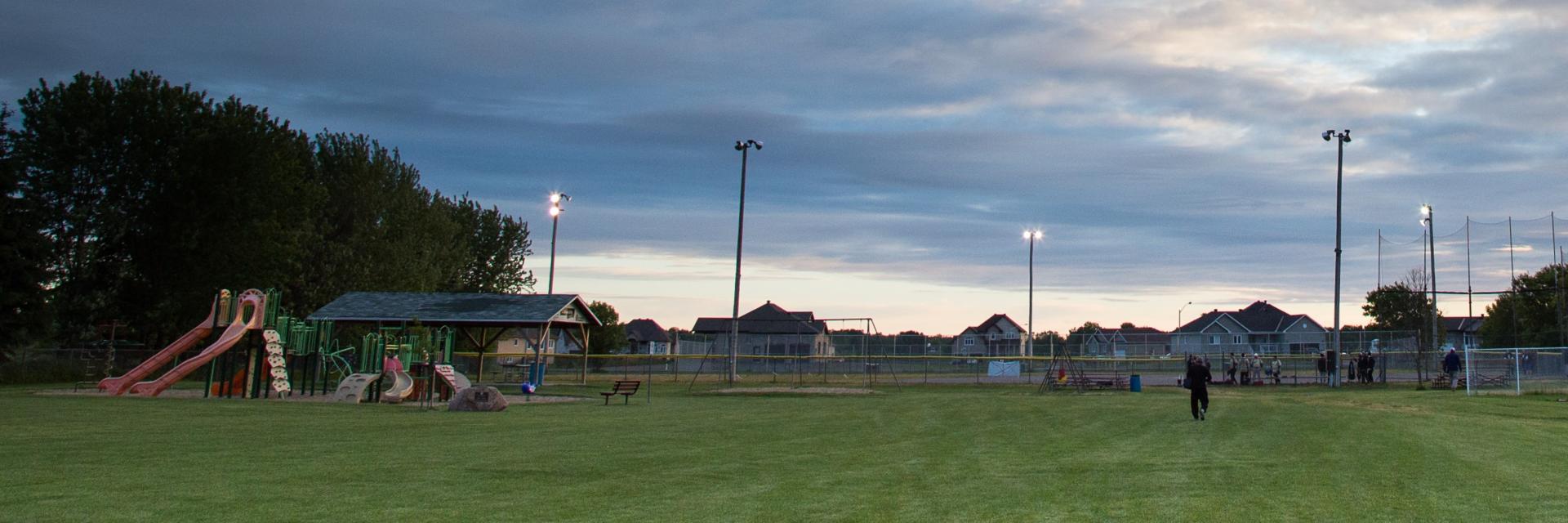 Playground at dusk with slide, trees, and light poles.