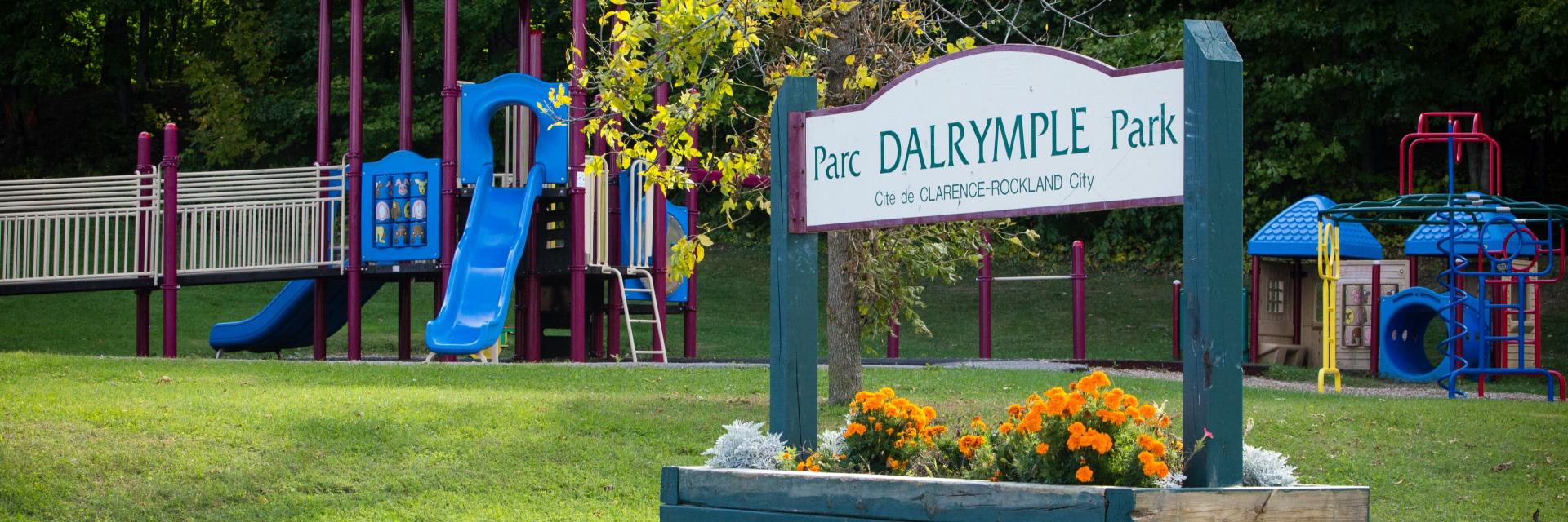 Playground with blue slides and sign, surrounded by green grass and trees.