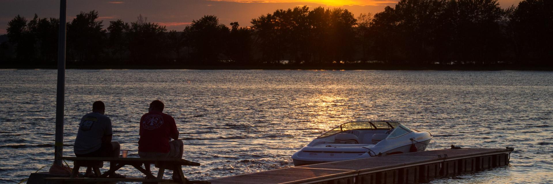 Sunset over a lake with two people seated on a dock, boat nearby.