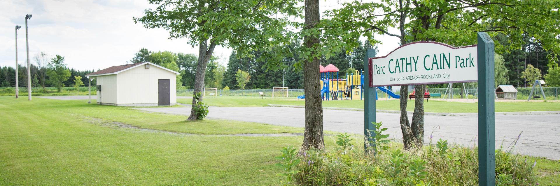 Park entrance with grassy area, trees, and small building in the background.