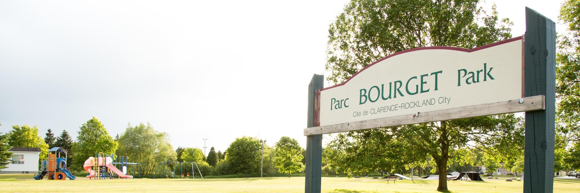 Park entrance sign with trees and playground in background.