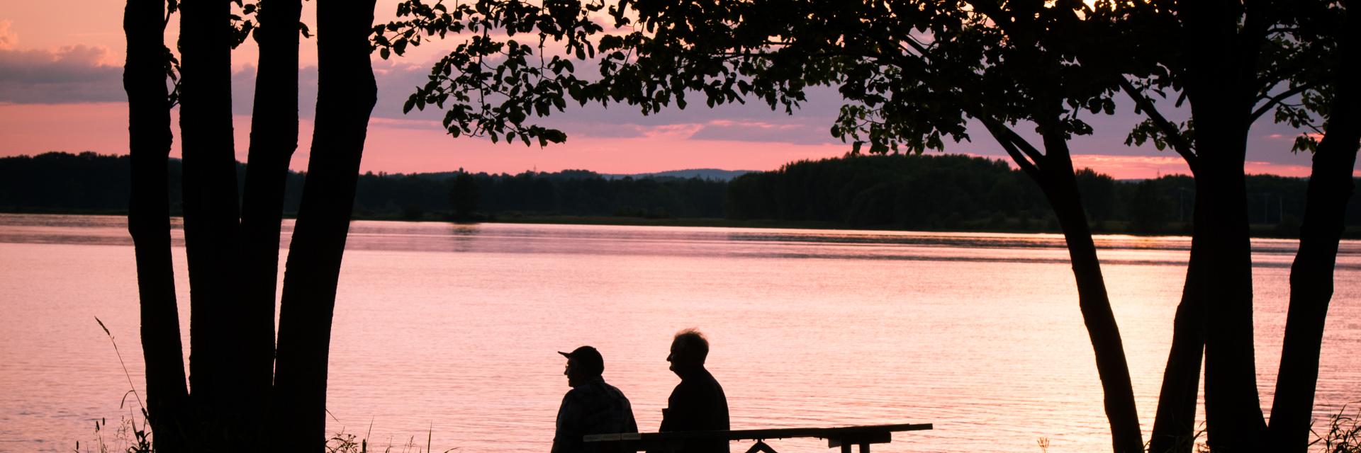 Two people seated at a lakeside picnic table during sunset.