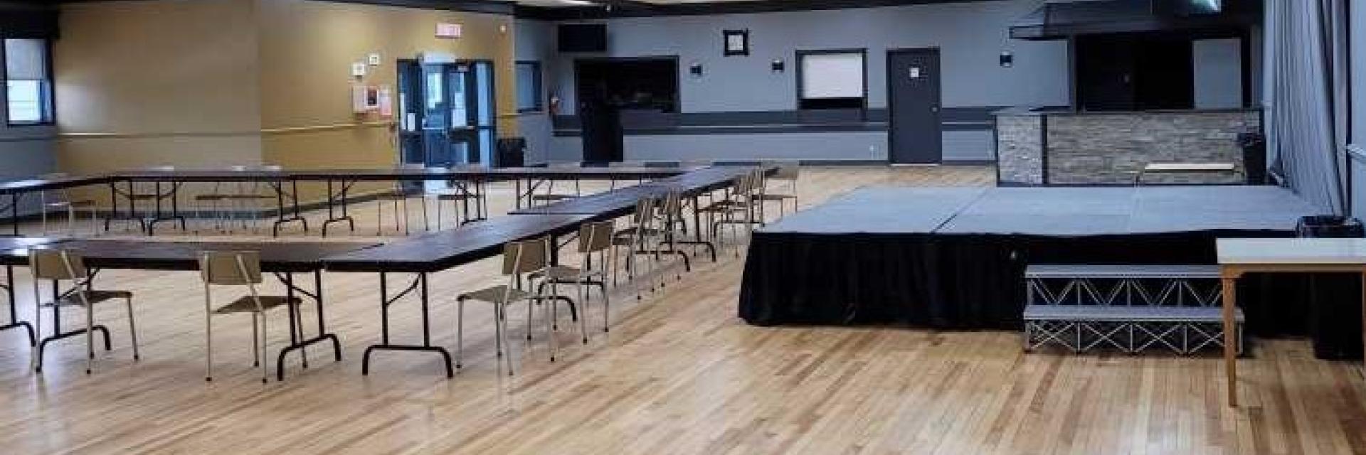 Spacious empty hall with wooden floors, tables, chairs, and a stage.