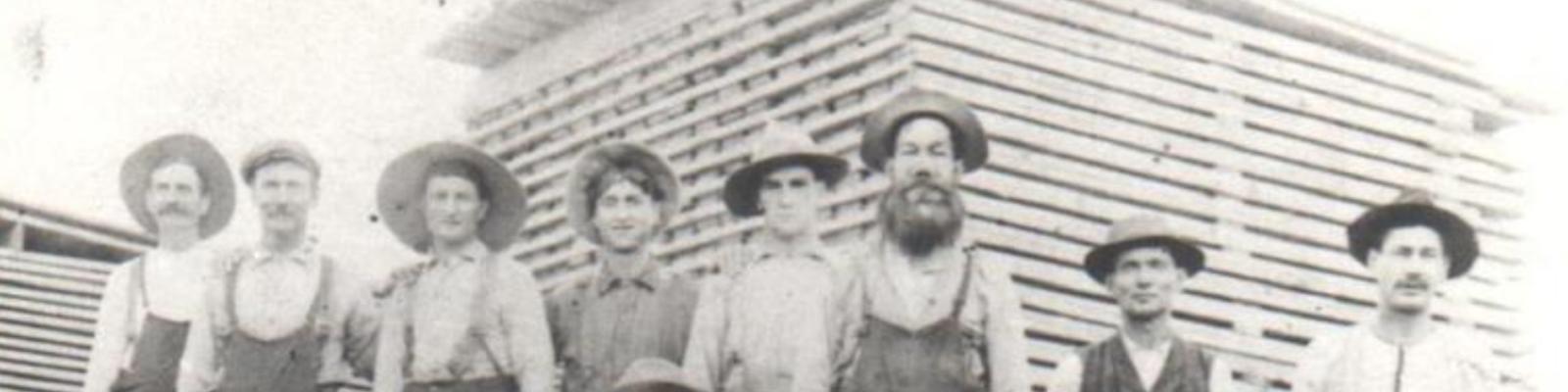 Group of people in hats standing in front of a wooden building, sepia-toned photograph.