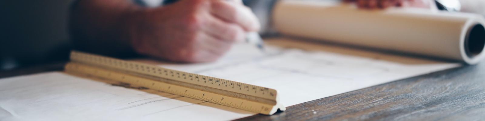 Person writing on architectural plans with a ruler on a table.