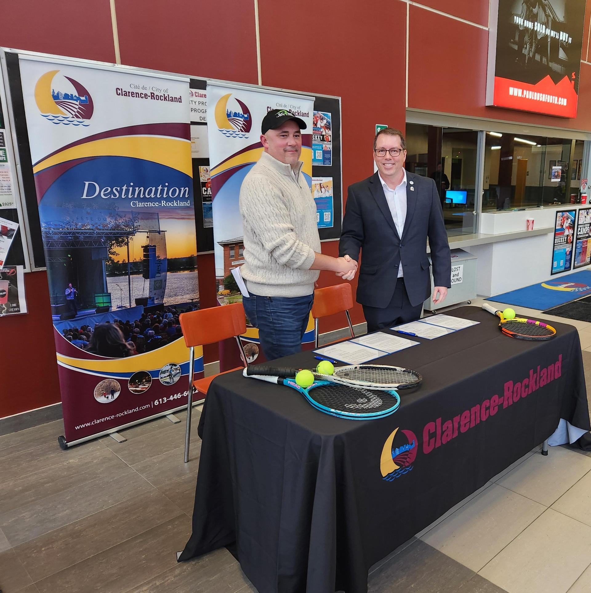Deux hommes se serrant la main à une table de signature avec des raquettes et des balles de tennis, devant des bannières promotionnelles de Clarence-Rockland